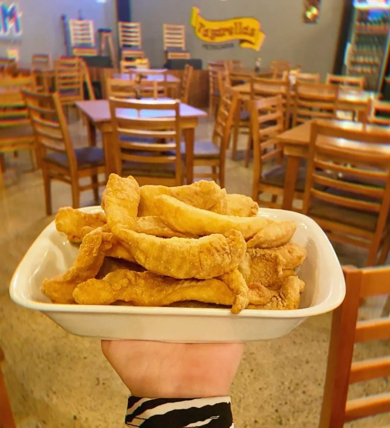 tagarelas-petiscaria-campo-largo- uma porção de peixe frito em uma bandeja e ao fundo muitas cadeiras em um salão amplo com o logo Tagarelas ao fundo